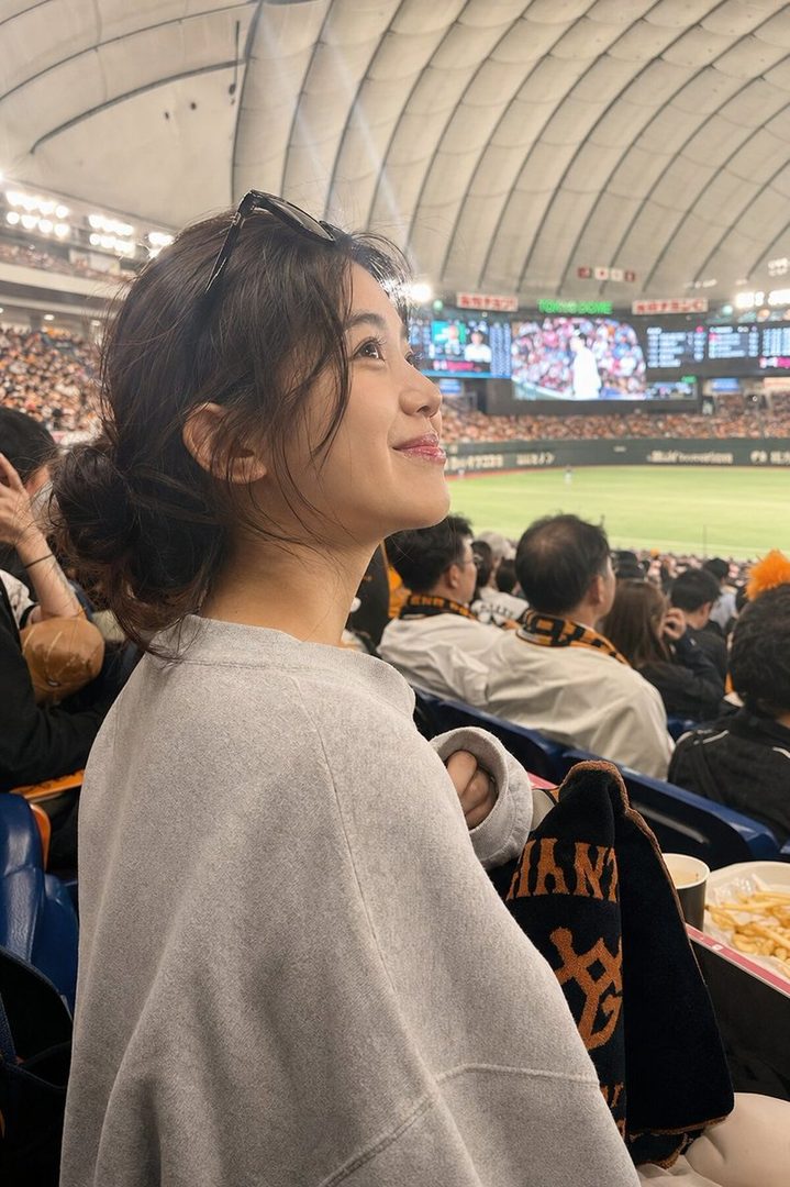 A young woman with dark hair in a messy bun, wearing a grey sweatshirt and sunglasses on her head, smiles while watching a baseball game in a stadium.