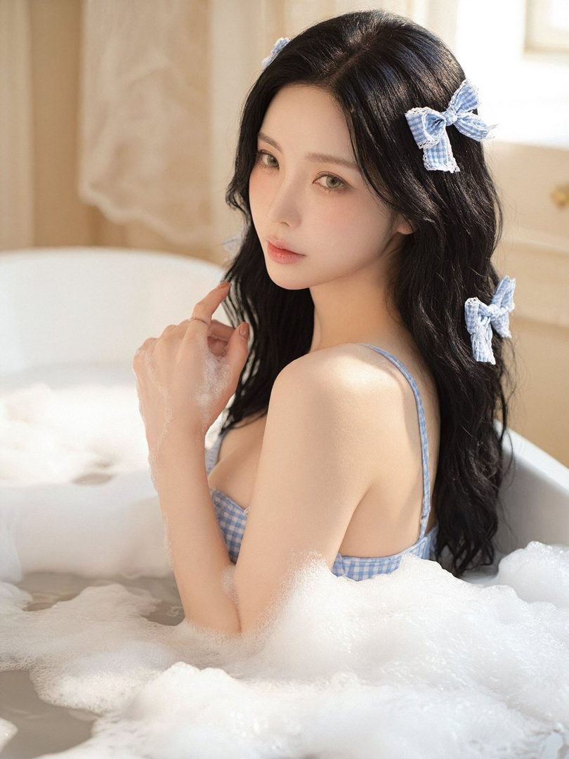A young woman with long black hair and blue gingham bows in her hair, posing in a bubble bath.