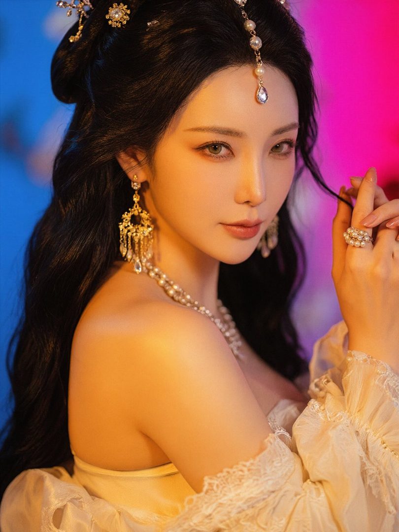 A beautiful East Asian woman with long black hair, adorned with intricate jewelry and a flowing white dress, poses elegantly in a studio setting with vibrant blue and pink lighting.