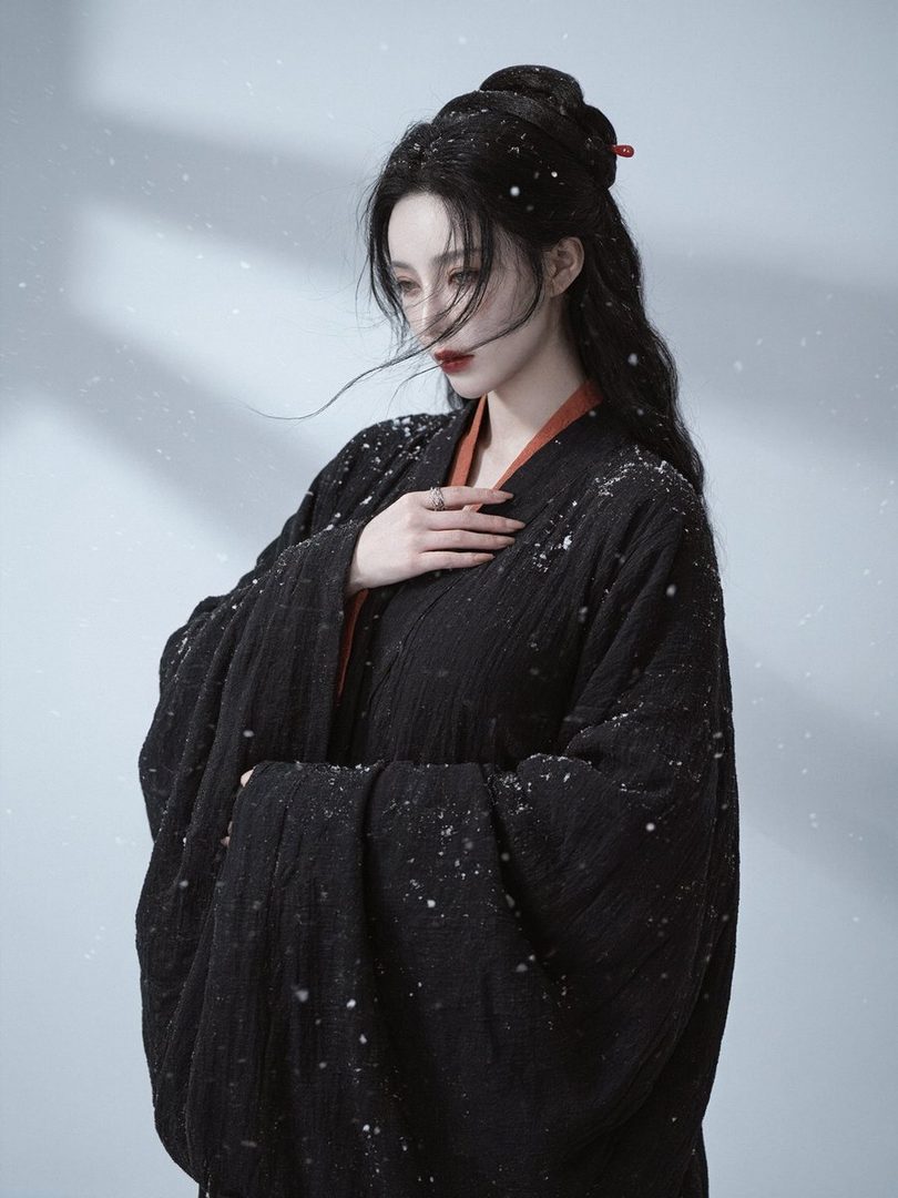 A woman with long black hair styled in an updo, wearing a black traditional robe with orange trim, stands in a snowy environment.