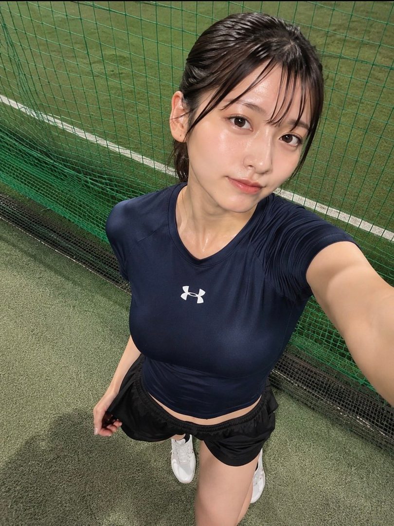 A young woman with wet hair tied back, wearing a navy blue athletic t-shirt and black shorts, poses for a selfie in an indoor sports facility.