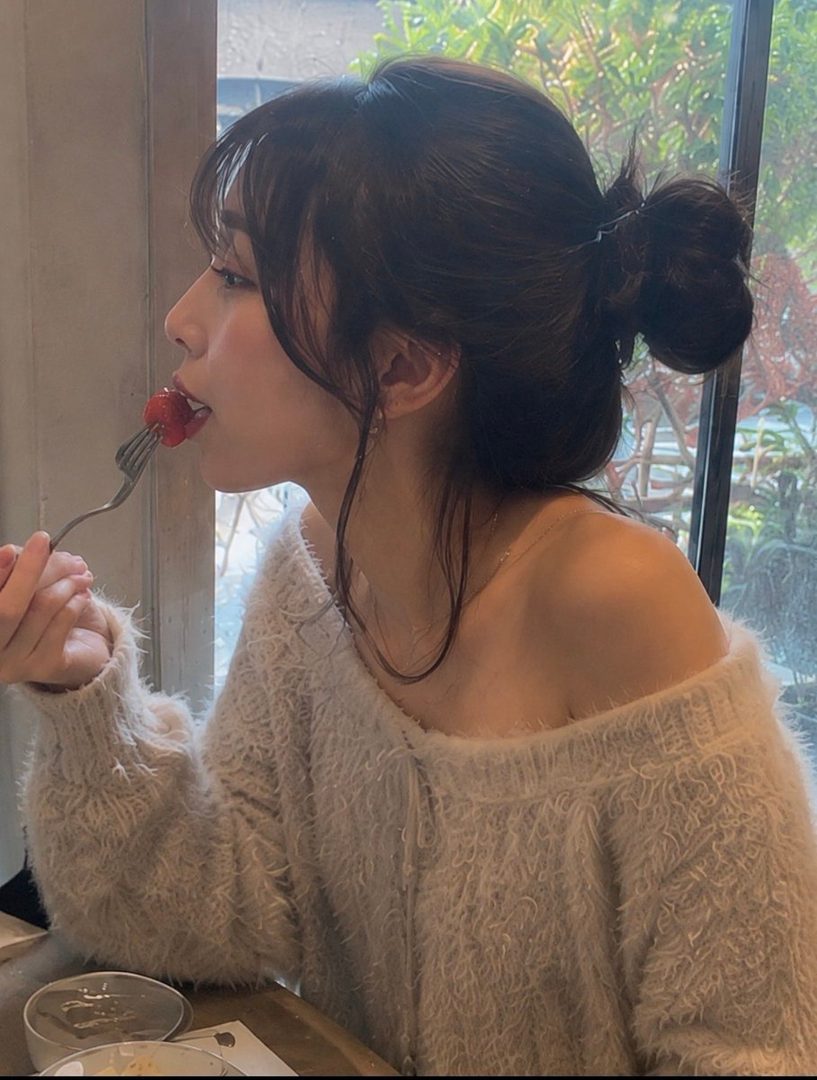 A young woman with her hair in a bun eats a strawberry with a fork, wearing a fluffy sweater.