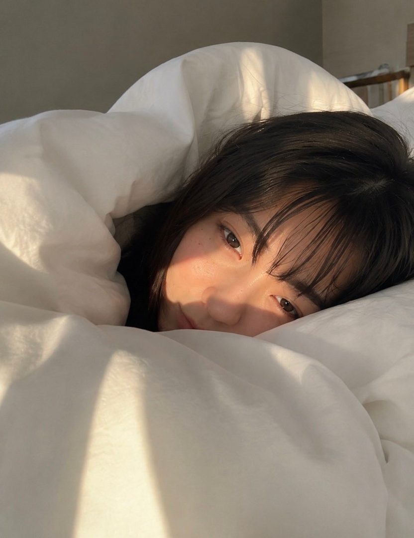 A young East Asian woman with dark hair and bangs, peeking out from under a white duvet in the morning sun, with a cute and relaxed expression.