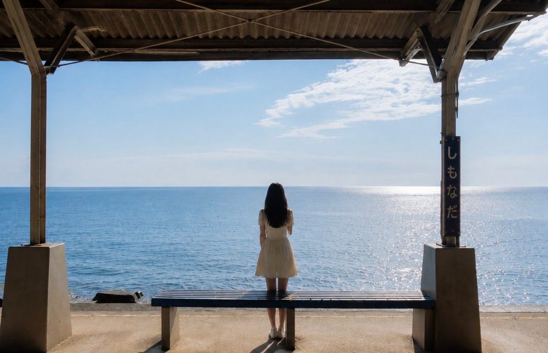 A young woman in a white dress stands on a platform overlooking the ocean, with a bench in front of her.