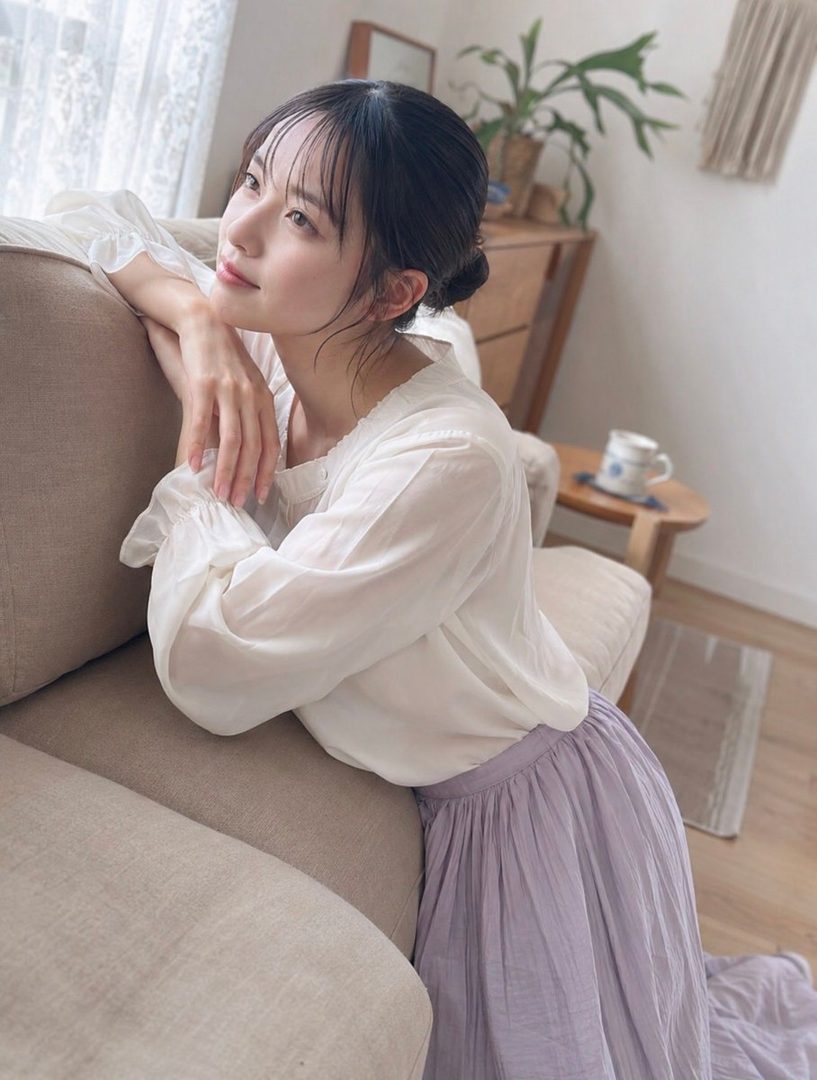 A young woman with black hair tied in a bun, wearing a white blouse and a light purple skirt, poses indoors by a sofa, looking thoughtfully out a window.