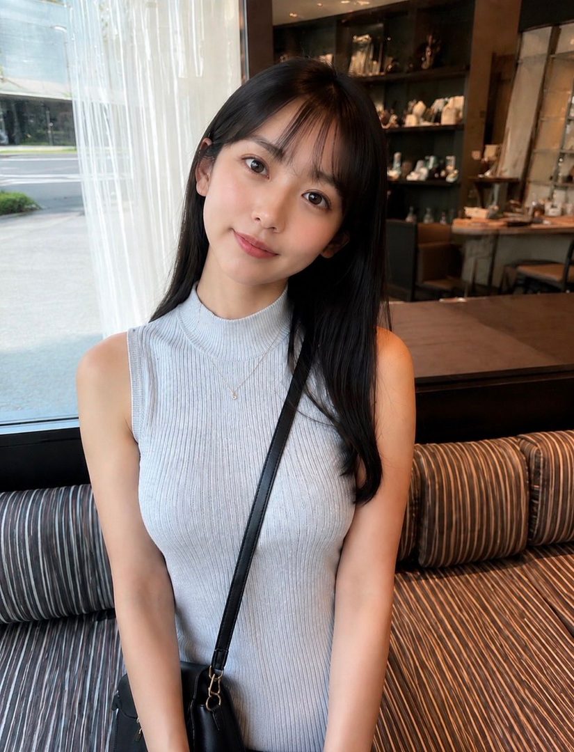 A young East Asian woman with long black hair and bangs, wearing a light blue sleeveless turtleneck top and a black crossbody bag, smiles gently while sitting indoors.