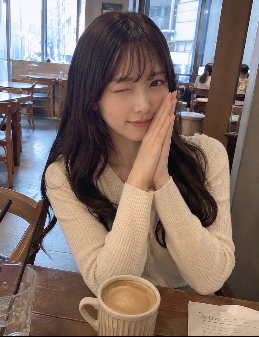 A young East Asian woman with long dark hair and bangs winks at the camera while resting her hands near her face, sitting at a table in a cafe with a cup of coffee in front of her.