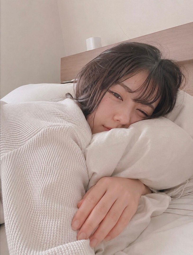 A young woman with short dark hair winks while lying in bed, wrapped in a white robe and blankets, with a soft morning light.