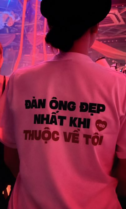A person wearing a white t-shirt with Vietnamese text and a black hat, seen from behind in a dimly lit, pink-hued environment.