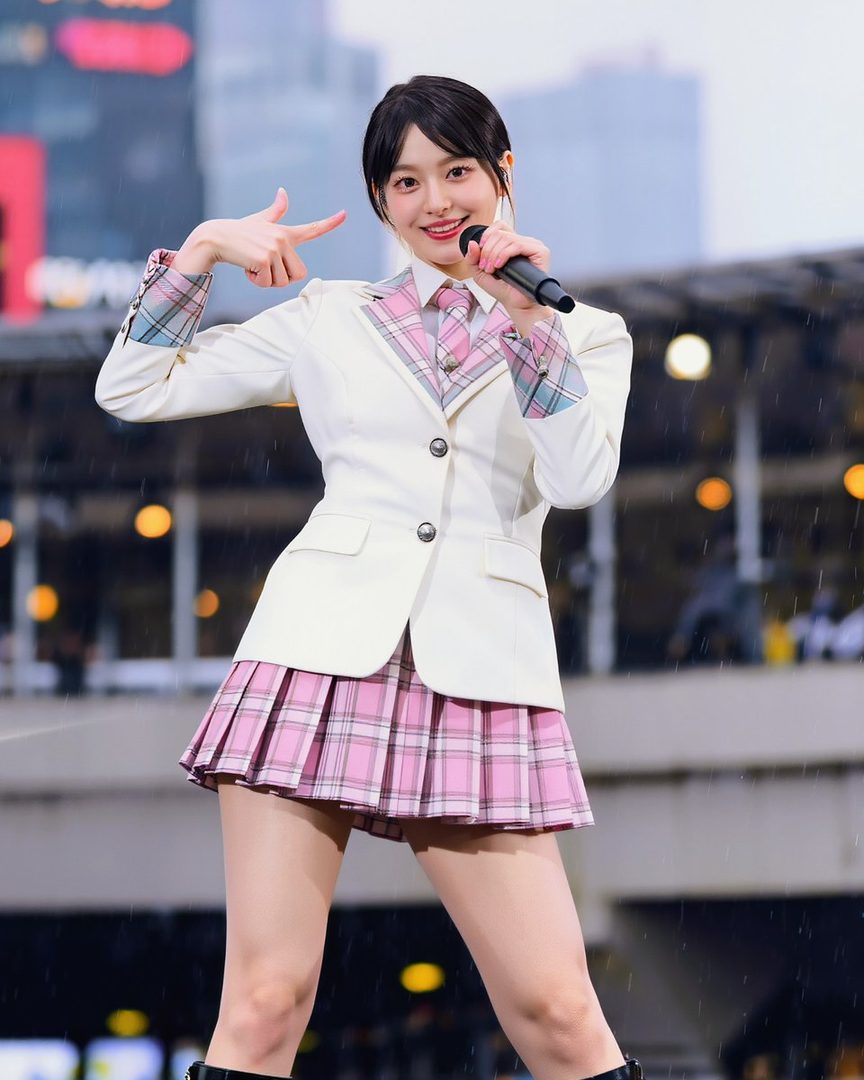 An AI idol in a white blazer, pink plaid tie, and pink plaid pleated skirt, holding a microphone and posing.