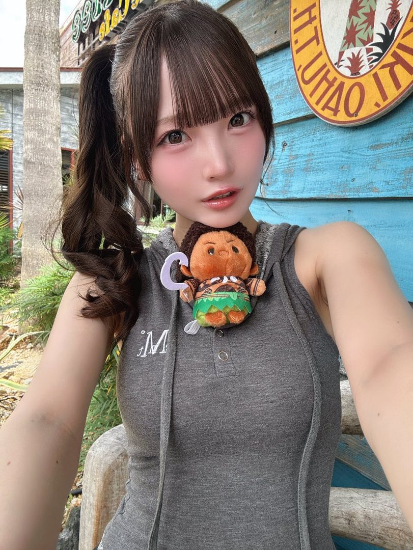 A young East Asian woman with brown hair in a ponytail takes a selfie outdoors, wearing a grey sleeveless top and a small plush toy on her chest.