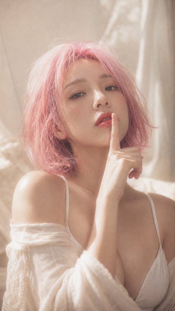 A young woman with short pink hair poses indoors, wearing a white top and looking at the camera with a finger to her lips.