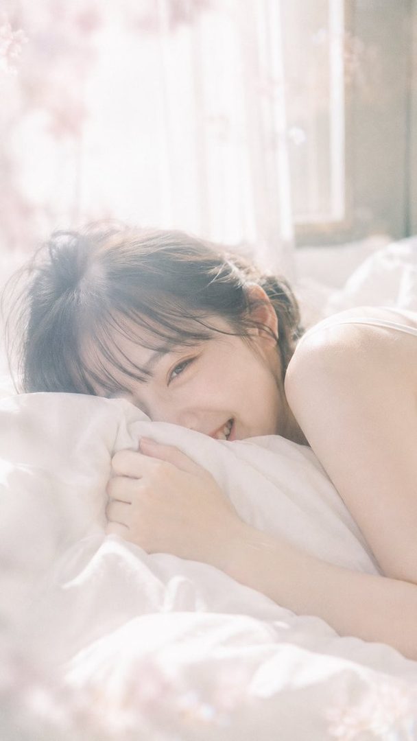 A young Japanese woman with dark hair, lying in bed with a pillow, smiling shyly in soft morning light.