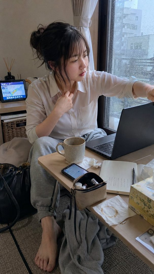 A young woman with her hair in a messy bun, wearing a white shirt and grey sweatpants, works on her laptop by a window on a rainy day.