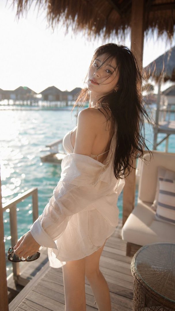 A woman in a white shirt and bikini stands on a wooden deck by the ocean, looking back over her shoulder.