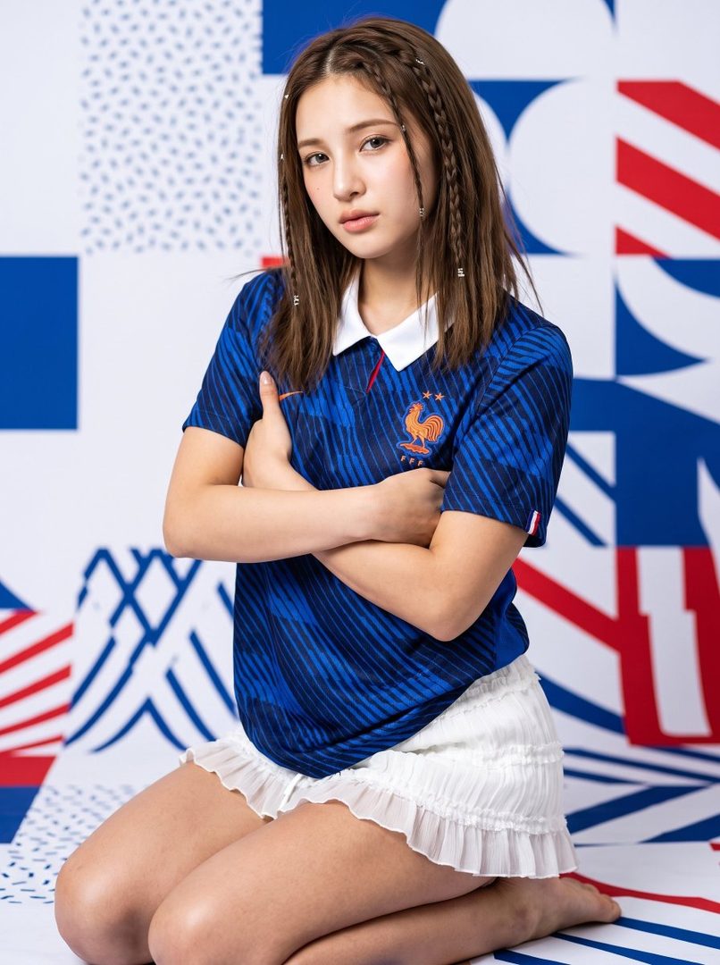 A young woman wearing a blue France national football team jersey and a white skirt, posing with her arms crossed in a studio setting with a blue and white patterned background.
