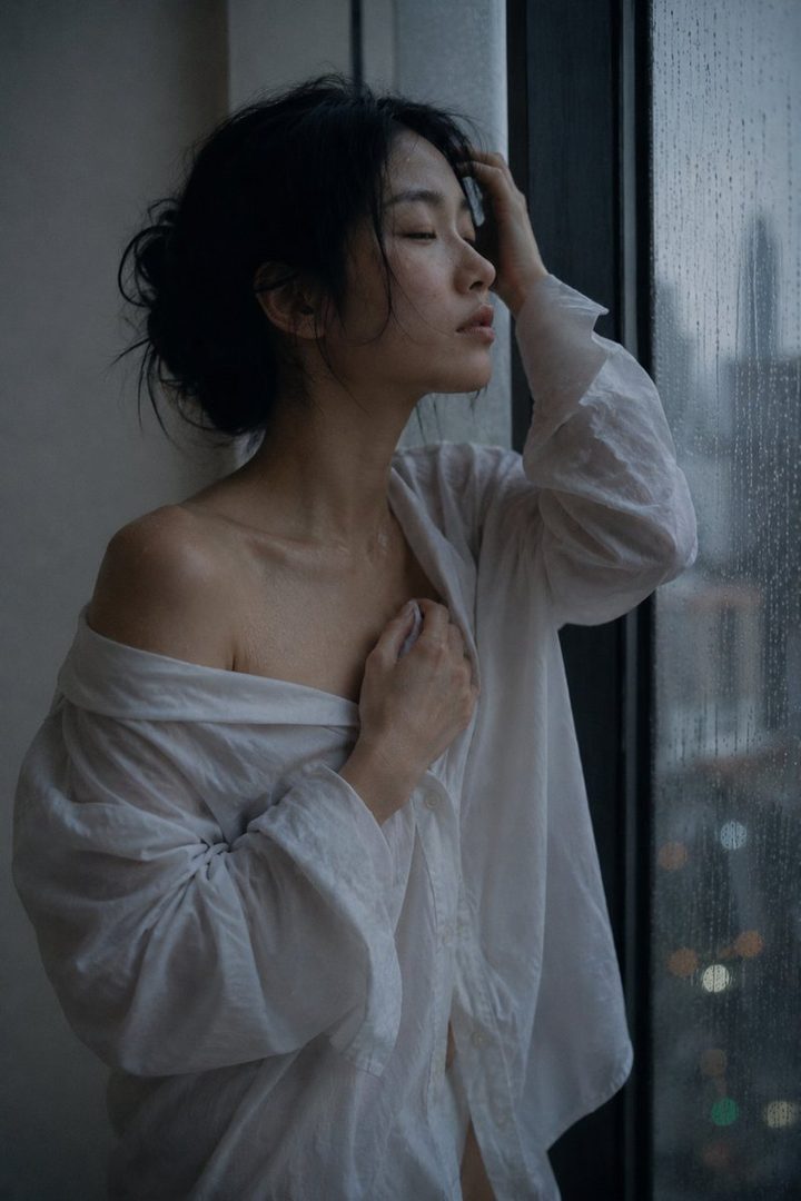A young East Asian woman in a white oversized shirt stands by a rain-streaked window, looking out pensively.