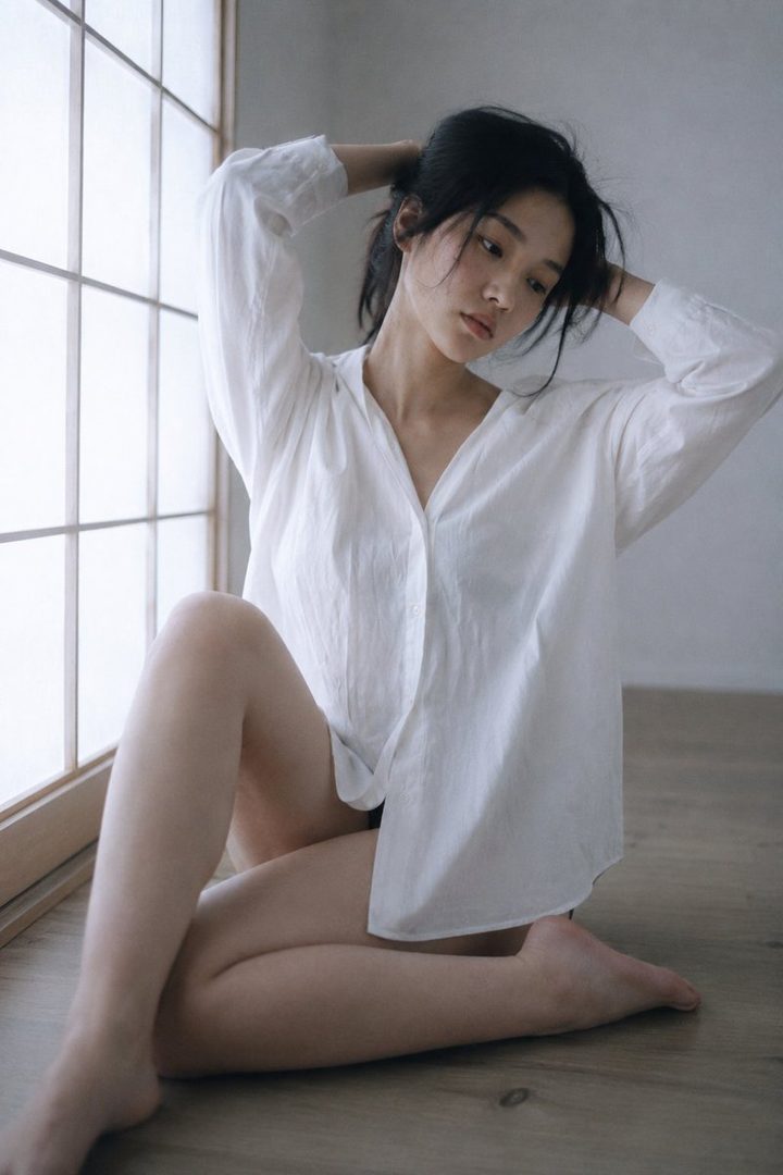 A young East Asian woman with black hair tied in a ponytail, wearing a white oversized shirt, poses indoors by a window with her hands in her hair.