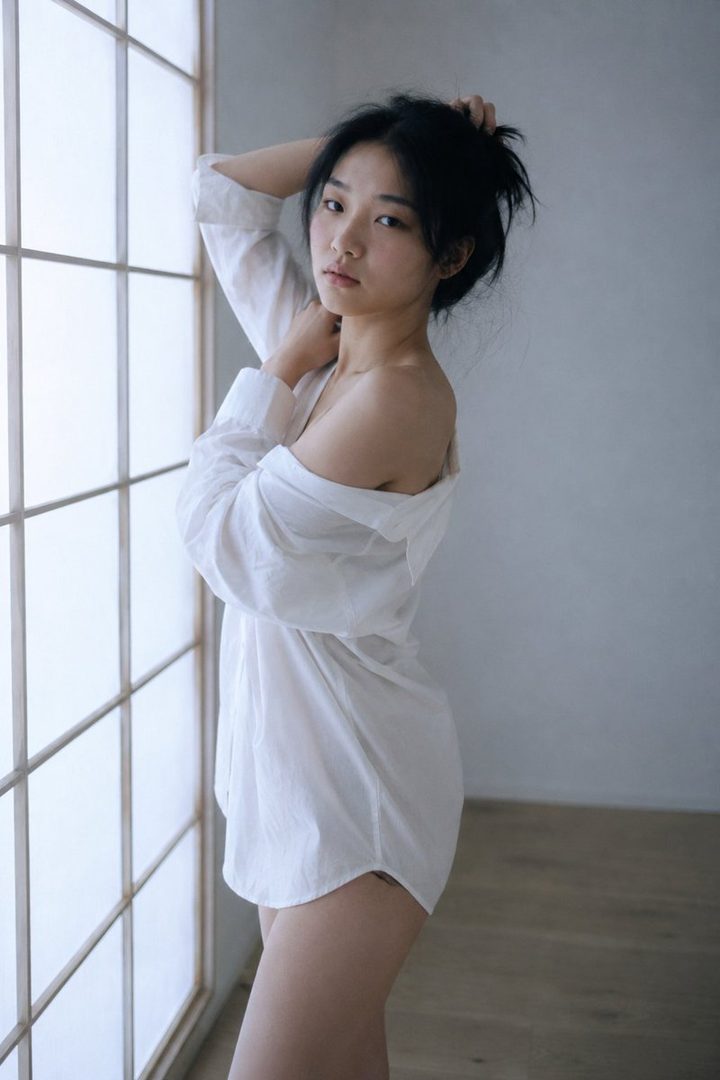 A young East Asian woman in an oversized white shirt poses by a window, her hair loosely gathered.