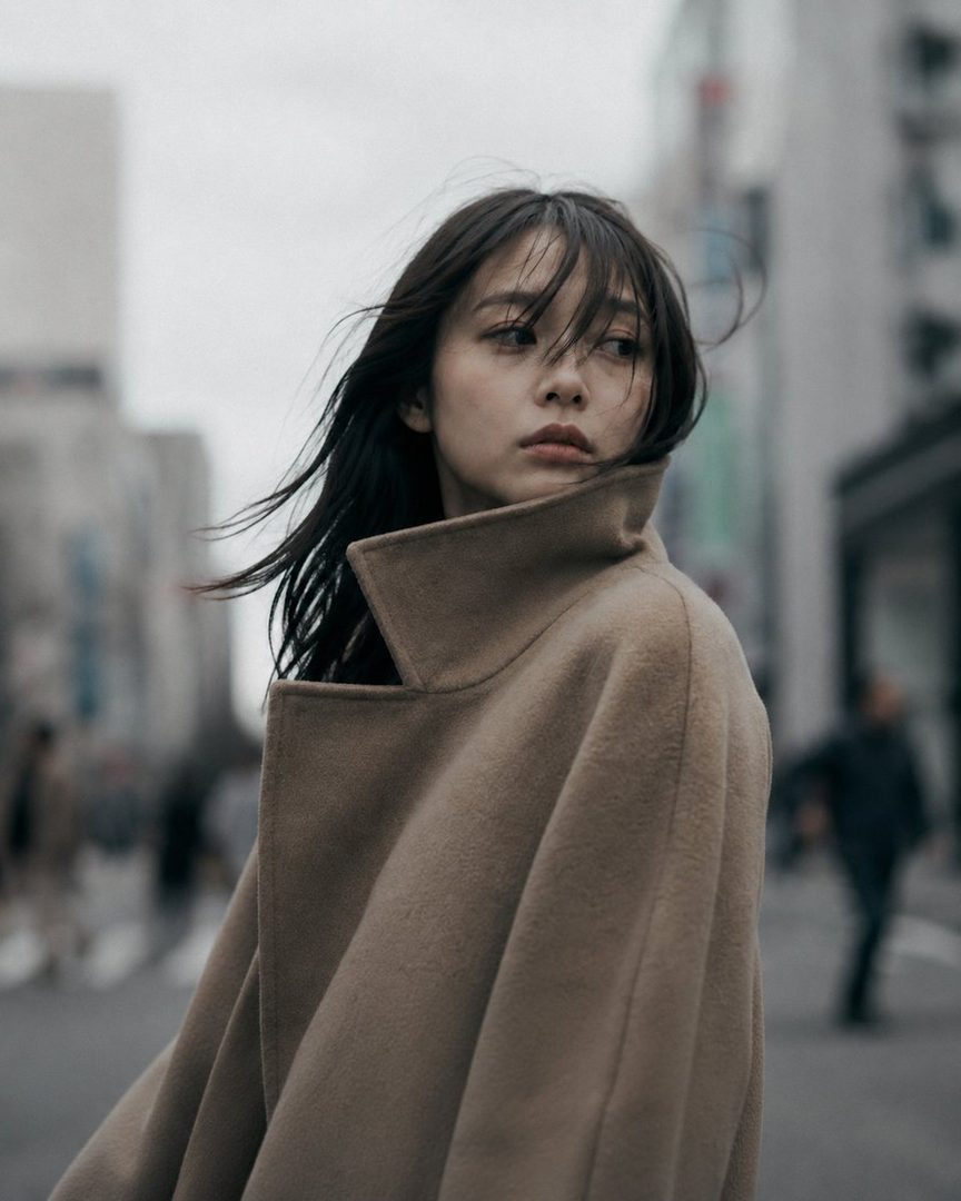 A Japanese woman in her mid-20s, wearing a camel beige wool coat with the collar up, stands on a blurred urban street with her dark hair blowing in the wind.