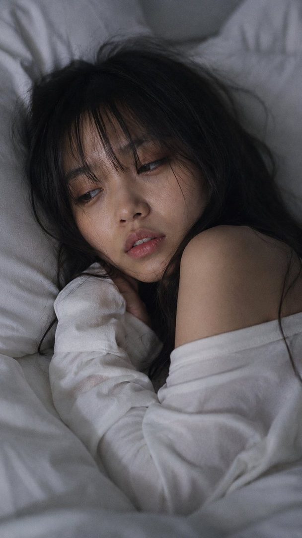 A close-up portrait of a young Japanese woman with long dark hair and bangs, lying in bed with her eyes half-lidded and lips slightly parted, wearing a white t-shirt.