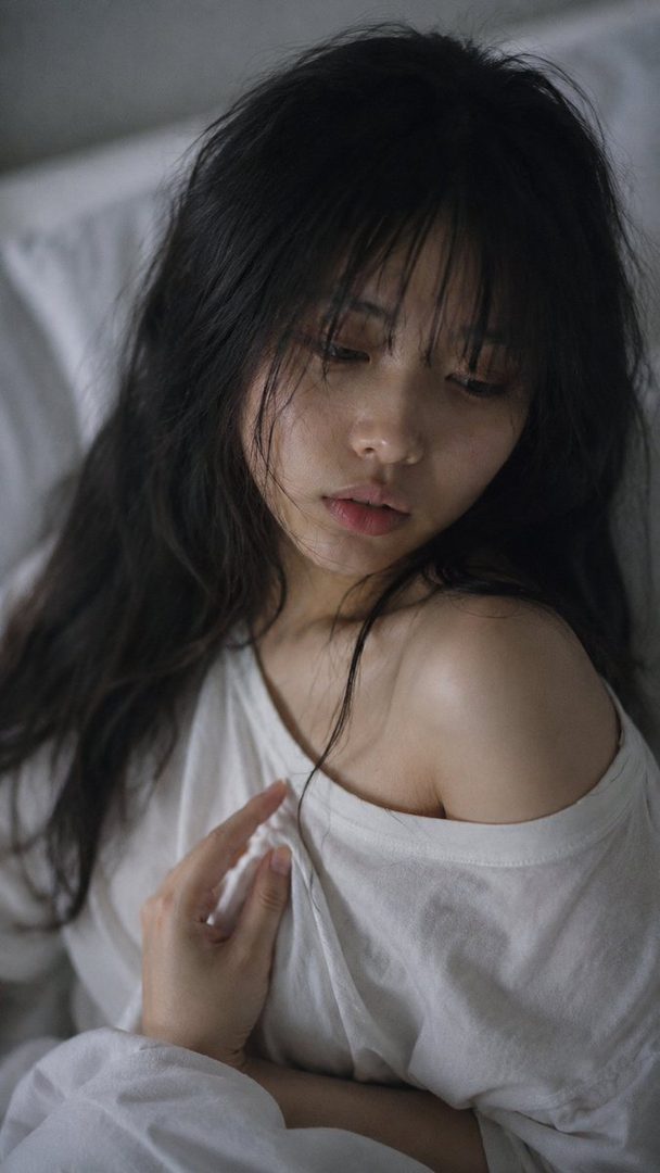 Close-up portrait of a young Japanese woman with long, dark, disheveled hair and bangs, wearing a white t-shirt with one shoulder exposed, looking drowsy in soft morning light.