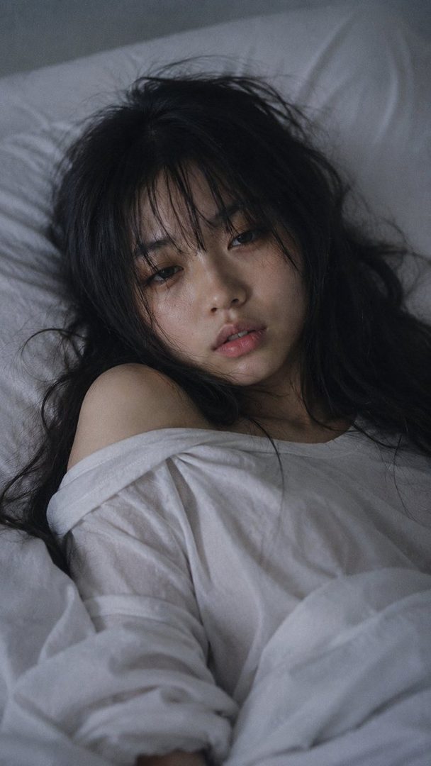 A close-up portrait of a young Japanese woman with long dark hair and bangs, lying in bed with her eyes half-lidded and lips slightly parted, wearing a white off-the-shoulder t-shirt.