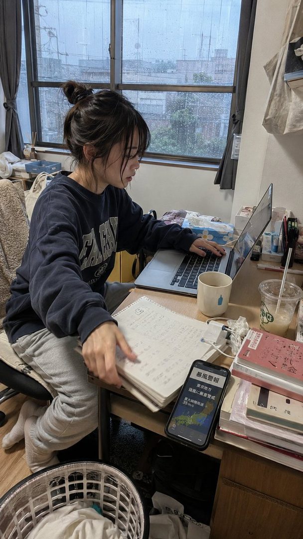 A young woman with dark hair in a messy bun and bangs is sitting at a desk, wearing a dark blue sweatshirt and grey sweatpants, looking at a notebook while a laptop is open beside her, with a rainy window in the background.