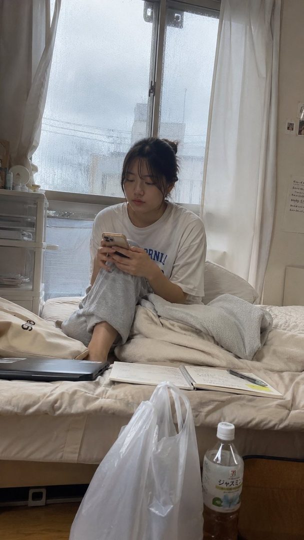 A young woman with dark hair in a messy bun sits on a bed, looking at her phone, with a rainy window in the background.