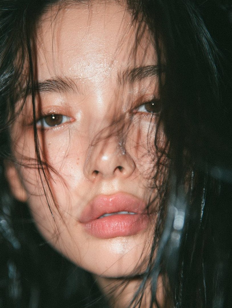 Close-up portrait of a young woman with dark, wet hair obscuring her face, soft lighting, and a dreamy expression.