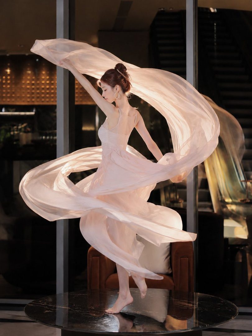 A graceful woman in a flowing pink dress dances with a sheer fabric, her arms outstretched.