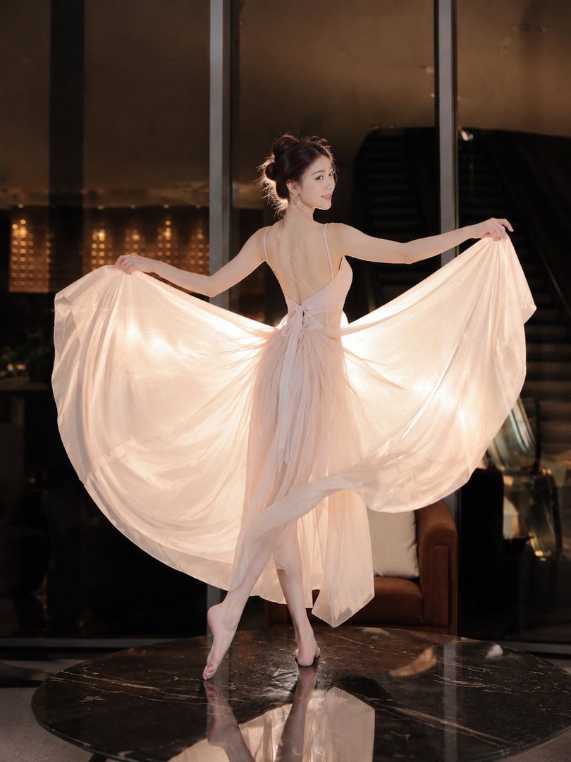 A graceful woman in a flowing pink dress poses elegantly indoors, her arms outstretched and her dress billowing around her.