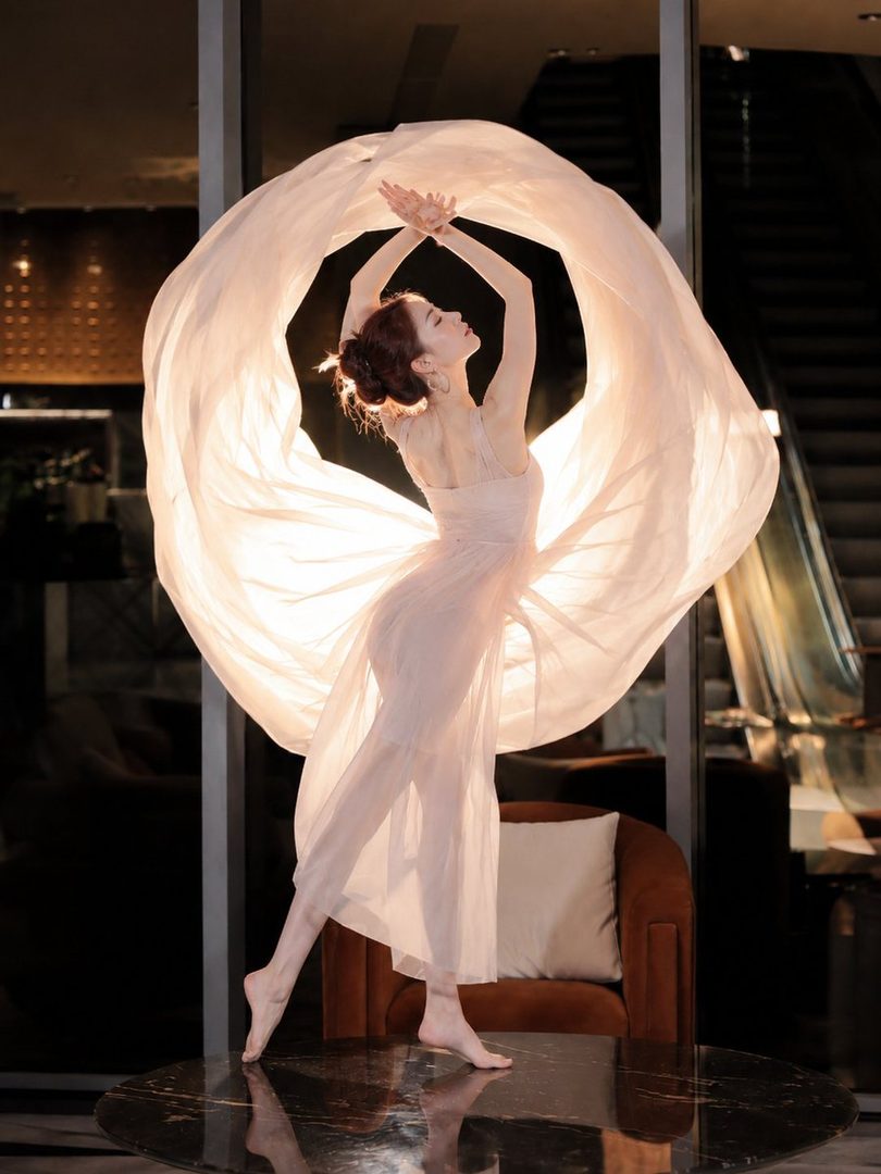 A graceful woman in a flowing light pink dress dances with a large sheer fabric, her arms raised and hands intertwined, against a dark, modern indoor background.