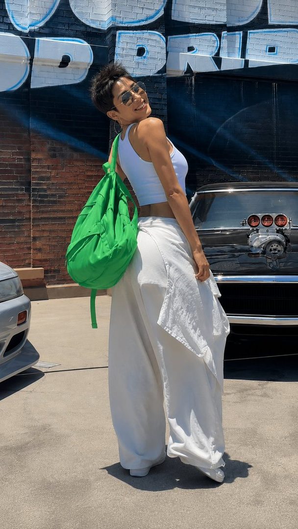 A woman with short dark hair and sunglasses smiles while wearing a white tank top and white wide-leg pants, carrying a green backpack, standing in front of a brick wall with graffiti and a car.