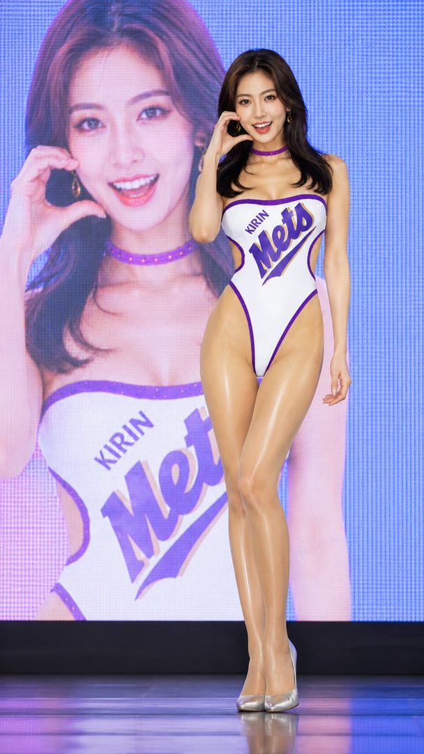 A beautiful East Asian woman in a white and purple high-cut swimsuit poses confidently, with a large screen displaying her image behind her.