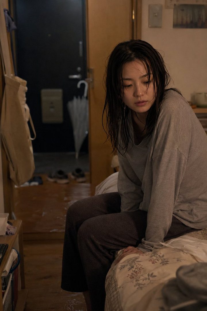 A tired woman with wet dark hair sits on a bed in a dimly lit room after coming home on a rainy day.