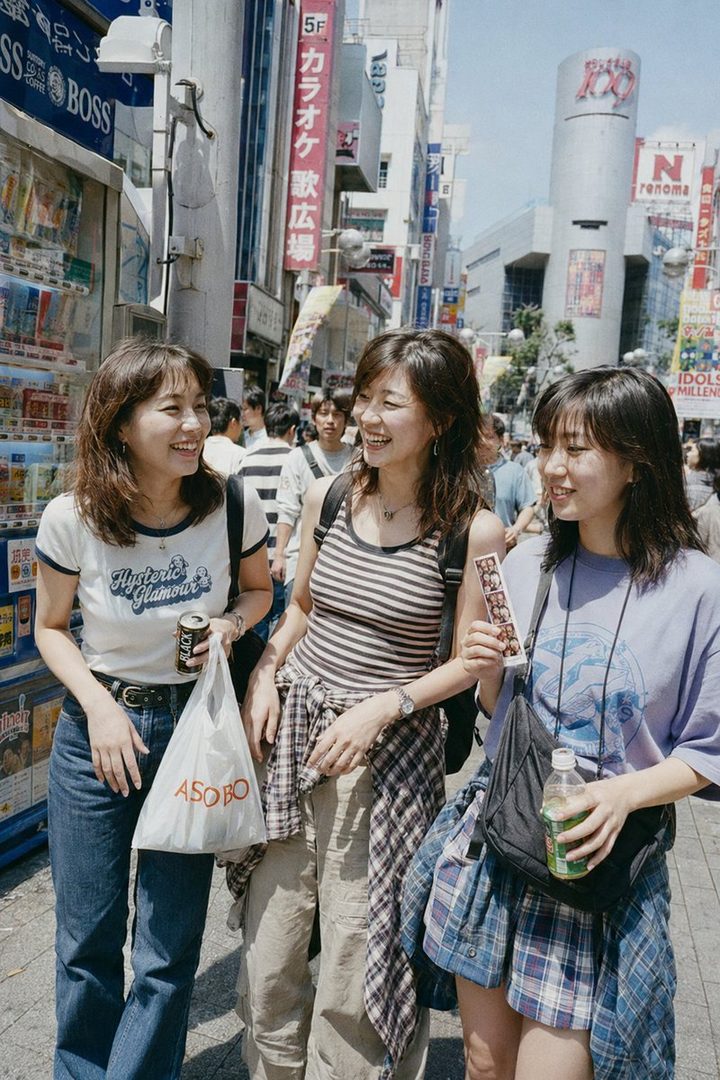 Three young Japanese women are laughing and walking together on a busy street in Shibuya, Tokyo, reminiscent of 1990s street photography.
