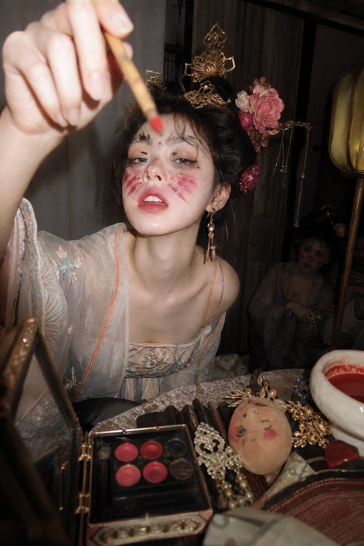 A young woman with elaborate traditional East Asian makeup and hair ornaments applies makeup with a brush, looking at the camera.