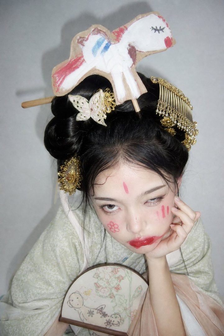 A young East Asian woman with elaborate traditional hairstyle and makeup, wearing a light green traditional outfit, holds a fan and poses playfully.