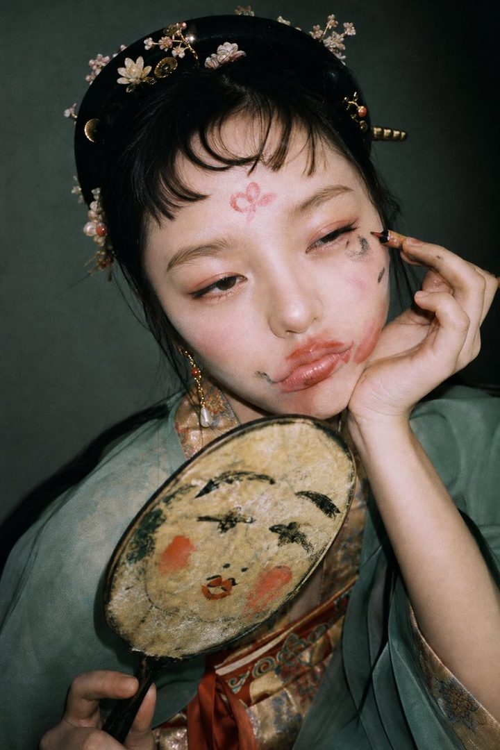 A young East Asian woman with dramatic makeup and traditional Chinese attire holds an antique hand mirror, looking thoughtfully to the side.