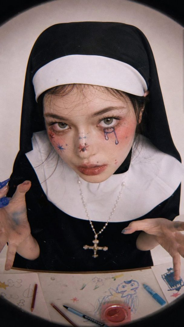 A young woman with striking makeup, dressed as a nun, poses with a playful and slightly unsettling expression, surrounded by children's drawings.