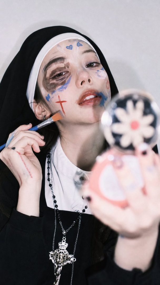 A young woman in a nun costume with artistic makeup, holding a makeup brush and a compact mirror.