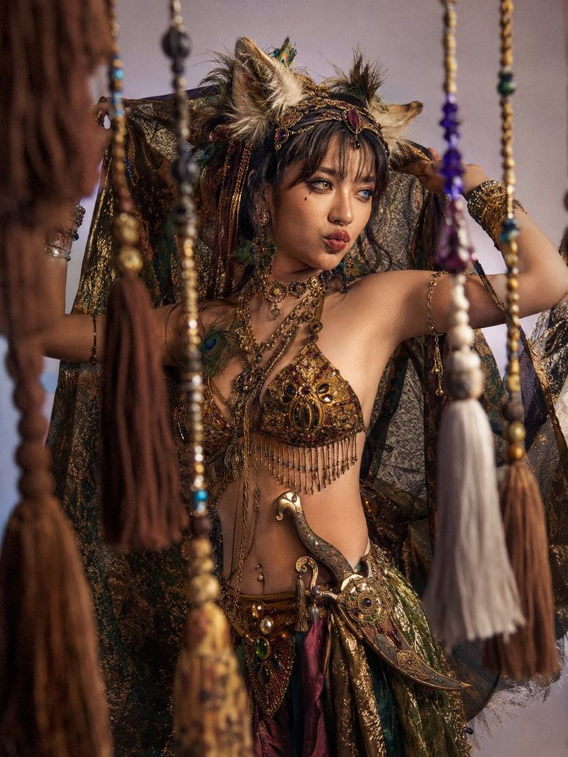 A woman in elaborate golden tribal costume with cat ears and peacock feather accents, posing playfully with a dagger.