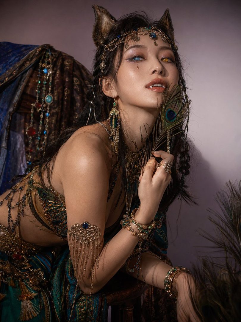 A woman with cat ears and heterochromia, adorned with peacock feathers and intricate jewelry, poses seductively.