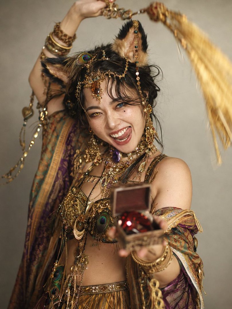 A playful woman with cat ears and elaborate golden jewelry, holding a small open box with a red gem, looking at the camera with a mischievous expression.