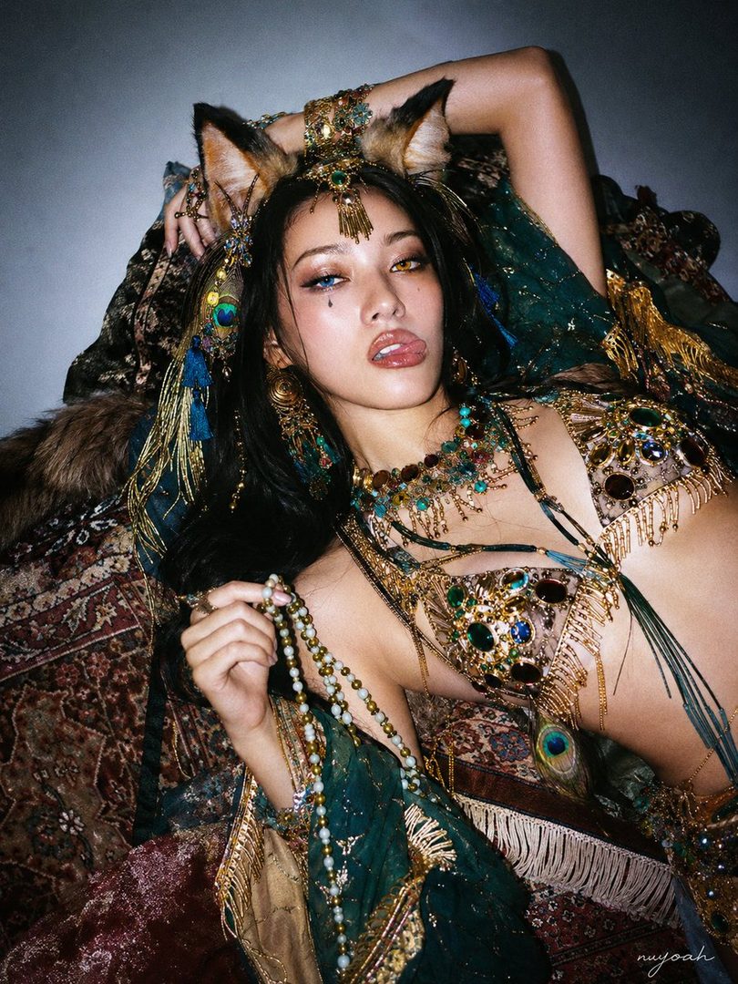 A woman with fox ears and heterochromia, adorned in ornate jewelry and a revealing outfit, lies on a patterned rug, holding a beaded necklace.