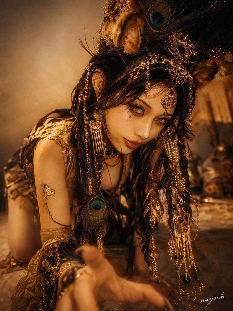 A woman with long dark hair adorned with peacock feathers and intricate gold jewelry, posing in a dramatic, fantasy-inspired outfit.