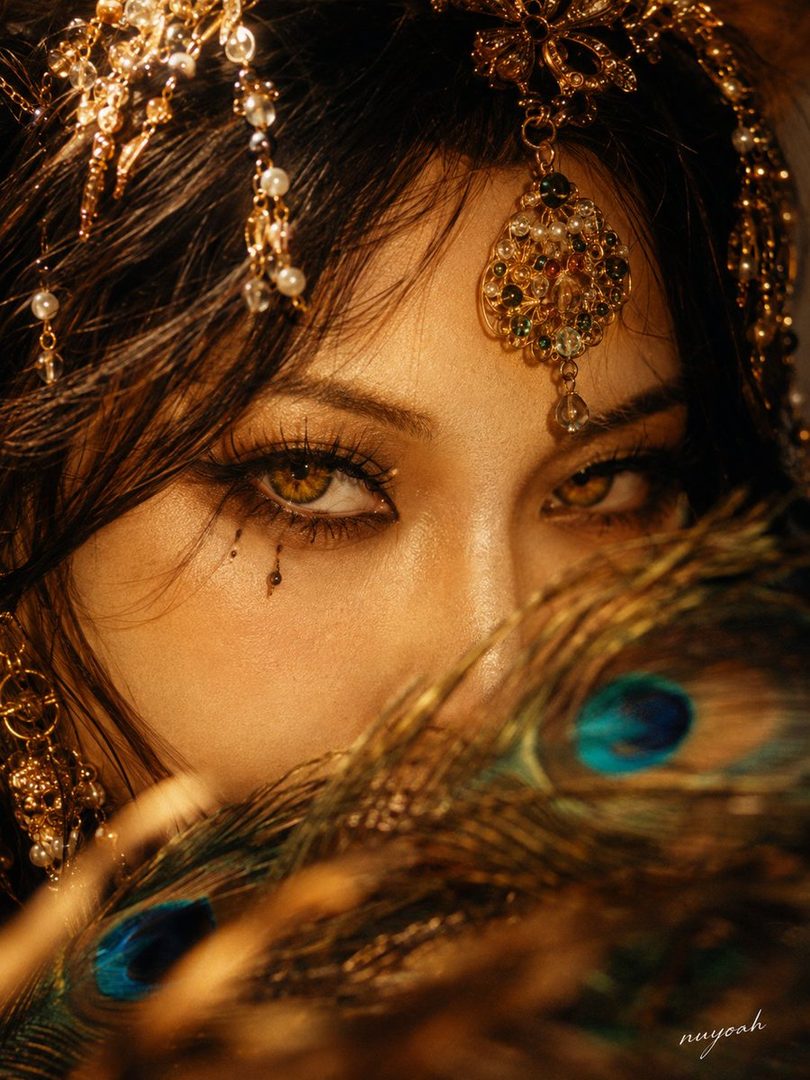 Close-up portrait of a woman with striking amber eyes, adorned with intricate gold jewelry and peacock feathers, evoking a mystical Persian-Bohemian fantasy theme.