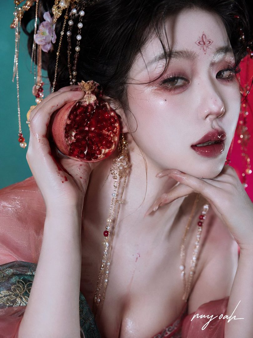 A woman with dark hair and elaborate traditional East Asian makeup holds a halved pomegranate, adorned with pearls and gold jewelry, in a suggestive pose.