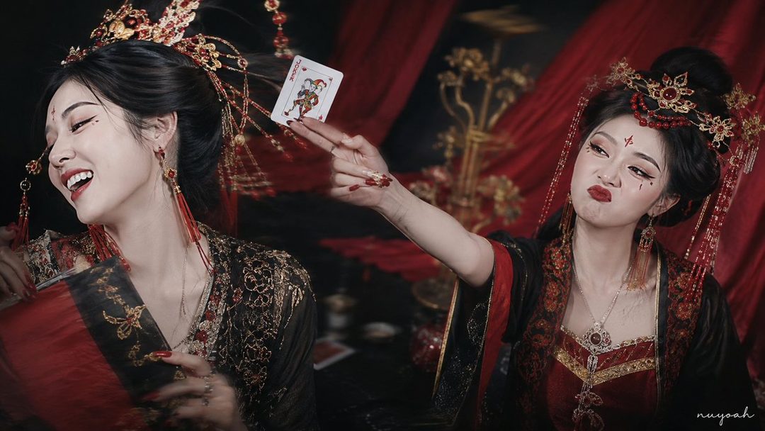 Two young women in traditional Chinese-inspired attire, one playfully holding a joker playing card, the other making a funny face.